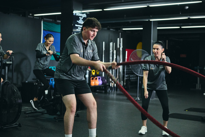 Participants in IPROWD training at a gym, with one person using battle ropes, another providing instruction, and others on exercise bikes, surrounded by kettlebells, weight racks, and a 'Strength' sign in the background.