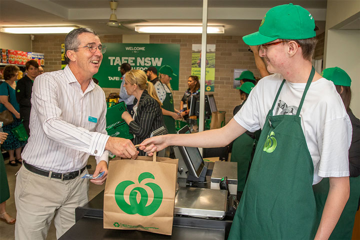 Student learning cashier skills in a Mini Woolies training environment, serving a customer at a checkout with other shoppers and staff in the background.