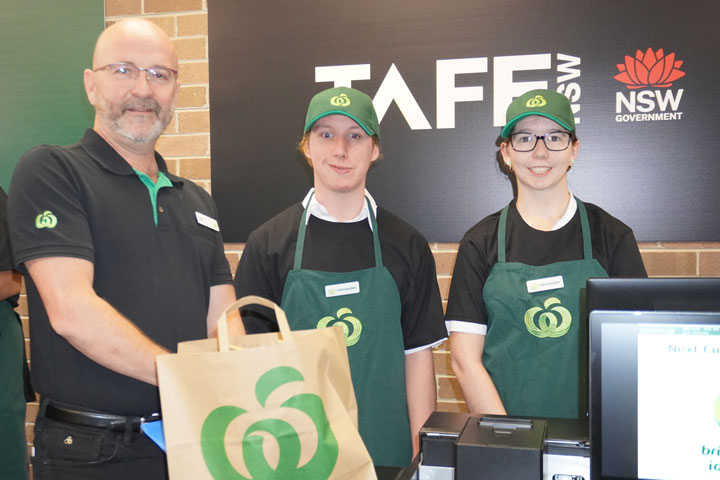 Three individuals in uniforms standing in front of a TAFE NSW sign, one holding a Woolworths branded tote bag.