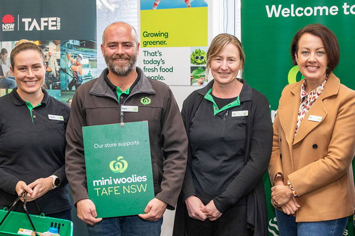 Four individuals standing side by side. They are wearing green and black uniforms with name tags, and one individual is holding a green folder with ‘Mini Woolies TAFE NSW’ written on it.