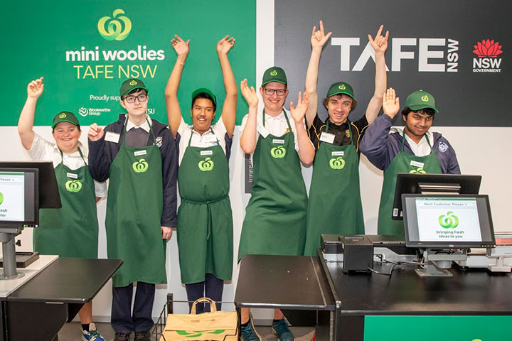Woolworths staff members standing at the checkout counter with Mini Woolies and TAFE NSW banners behind them