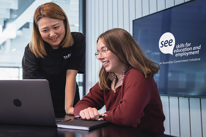 Two individuals in a professional setting, one using a laptop and the other writing notes, with a ‘SEE - Skills for Education and Employment’ logo on the screen in the background.