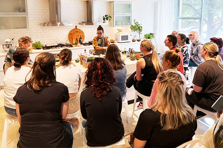 A group of women sit in a modern kitchen setting, watching two chefs demonstrate cooking techniques. The room is bright with natural light from large windows, and the chefs are surrounded by fresh herbs and ingredients. The kitchen features sleek white tiles, stainless steel appliances, and wooden chopping boards. The audience observes the chefs closely as they prepare food on a counter.