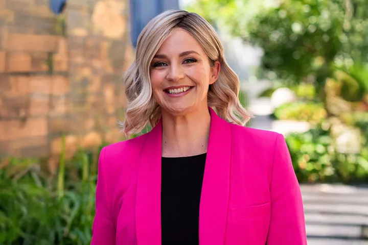 Amanda Rose stands outdoors wearing a bright pink blazer over a dark top, positioned along a garden walkway with greenery and stone walls in the background, promoting a Women in Business webinar focused on dealing with difficult people, protecting boundaries, and staying composed under pressure.