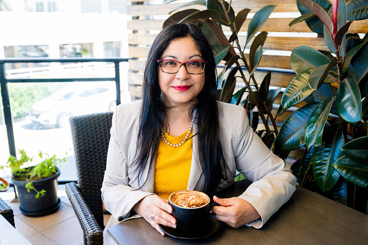 Dr Asheena Budhai Manuel sits at an outdoor café table wearing a light blazer and yellow top, holding a cup of coffee, with greenery, wicker seating, and natural timber screens in the background, promoting the Women in Business webinar on harnessing the power of self‑motivation.