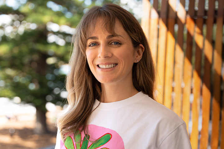 Laura Prael, Women in Business webinar presenter on '10 Years, 10 Lessons – A Founder’s Diary,' standing outdoors in front of trees and wooden panels, wearing a white t-shirt with a colorful graphic design.