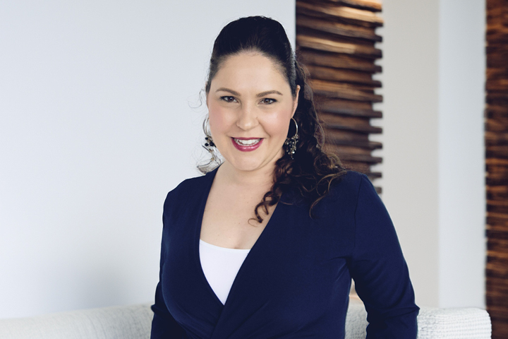 A person wearing a dark blue blazer over a white top, standing in front of a white wall with wooden slats on the right side.