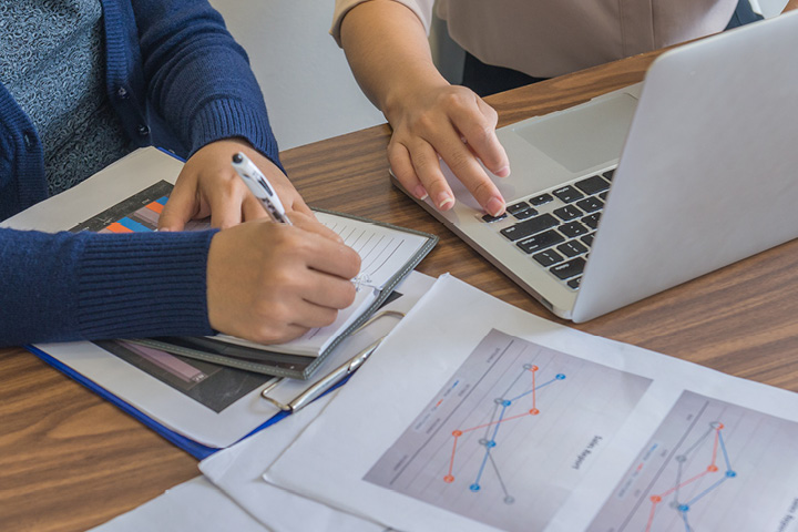 Close‑up of two individuals working at a table on personal finance planning, with one person writing notes beside printed charts and the other using a laptop to review financial graphs.