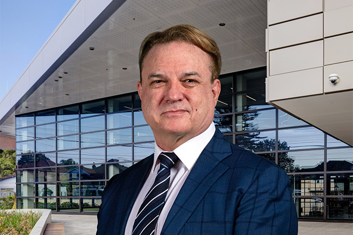Person wearing a navy suit and striped tie stands in front of a modern glass‑fronted TAFE NSW campus building with large windows, metal cladding, and landscaped surroundings.