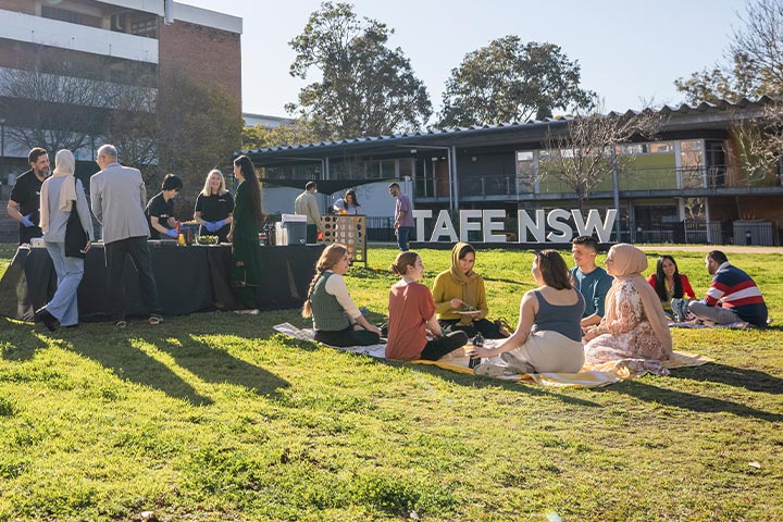 Group of people socialising outdoors on a sunny day near a TAFE NSW campus building, with tables set up and small groups seated on the grass.