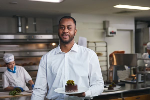 A man in a white collared shirt is holding a plate with a chocolate cake on it. He is in a TAFE NSW commercial kitchen.