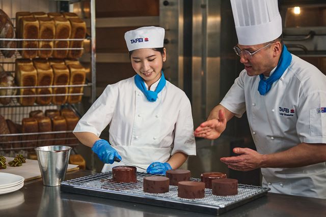 A TAFE NSW pastry student, guided by a professional chef, carefully decorates chocolate-coated desserts in a commercial baking kitchen.