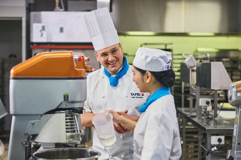 A professional chef in a white uniform and tall hat is smiling while guiding a student in a commercial kitchen. The student, also in a chef's uniform, is holding a measuring jug and engaging in the conversation.