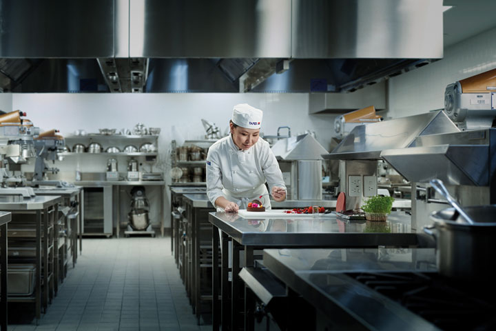 A professional chef in a TAFE NSW uniform is carefully plating a dessert in a commercial kitchen equipped with stainless steel appliances and workstations.