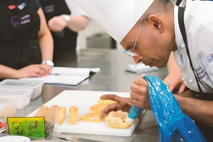 A professional chef is piping intricate designs onto pastries during a culinary class, while attentive participants observe in the background.