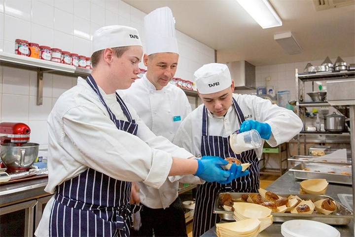 TAFE NSW patisserie students work with their teacher to sugar some baked goods. They are in TAFE NSW cookery uniforms.