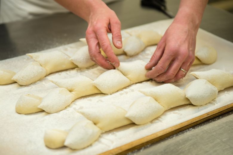 A baker’s hands carefully shape freshly prepared, flour-dusted dough into a decorative pattern on a wooden board.