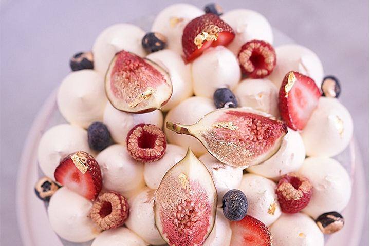 A beautifully decorated cake topped with piped cream, fresh figs, strawberries, raspberries, blueberries and gold leaf on a light pink cake stand.