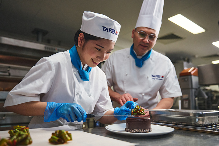 A TAFE NSW student decorates a chocolate dessert under the guidance of a chef instructor in a professional kitchen. Both are wearing chef uniforms and smiling as they work.