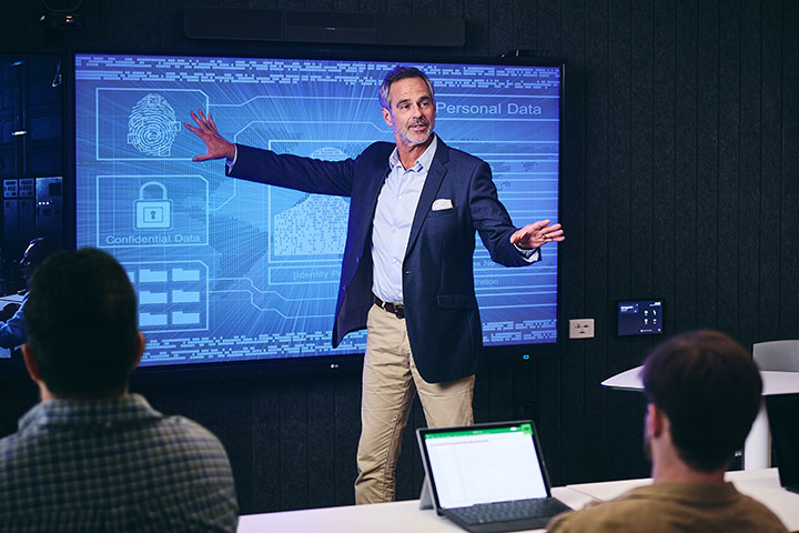 A well-dressed classroom facilitator stands beside a digital display featuring a visual schema and information data security.