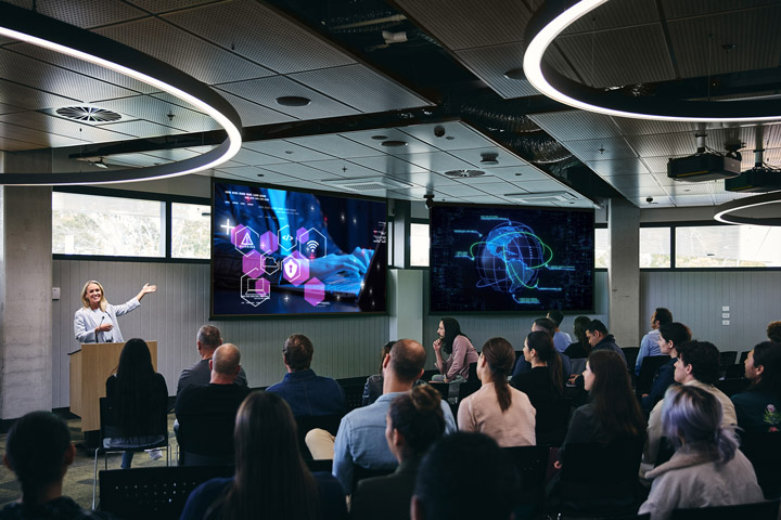 Digital teacher presents to a group of students seated in a modern lecture theatre, gesturing toward large wall‑mounted screens displaying data visualisations and connected technology concepts, within a contemporary IAT Digital learning space featuring circular overhead lighting.