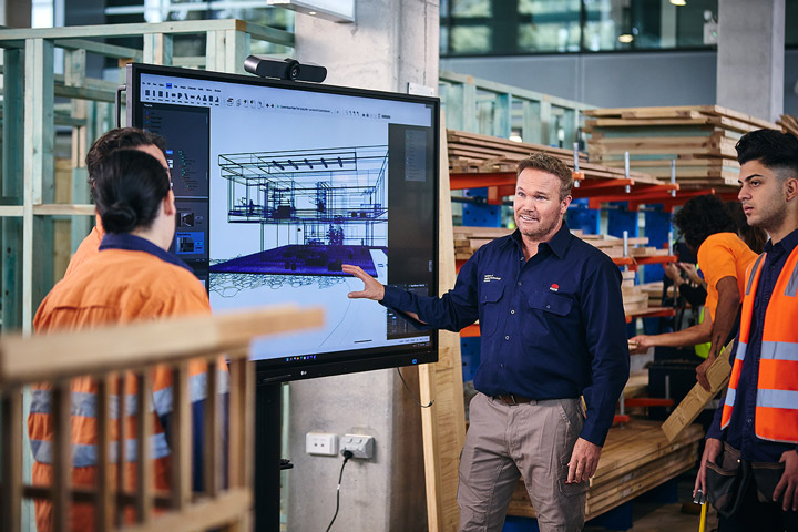Institute of Applied Technology Construction teacher explains a digital 3D building model on a large interactive screen to a group of students in a construction workshop, with timber framing, materials, and workstations visible behind them during a hands‑on learning session.