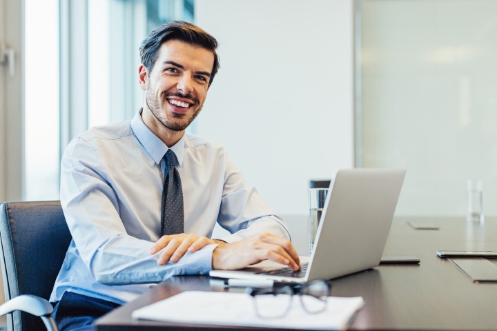 A man in a professional light blue shirt and tie sits at a desk working on his laptop. There is a notepad, pen and glasses in the foreground. He’s smiling at the camera.