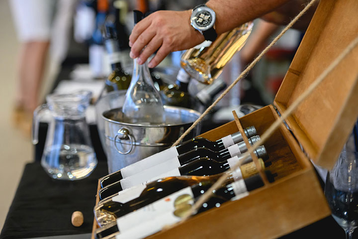 Wine bottles are decoratively displayed in a wooden box on a table. A hand nearby is placing a wine bottle in a stainless steel ice bucket.