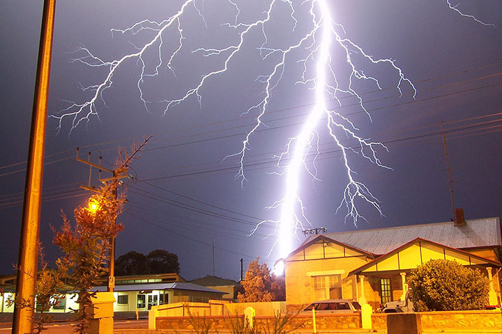 A massive bolt of lightning flashes in the night sky, illuminating the houses and suburb around it.