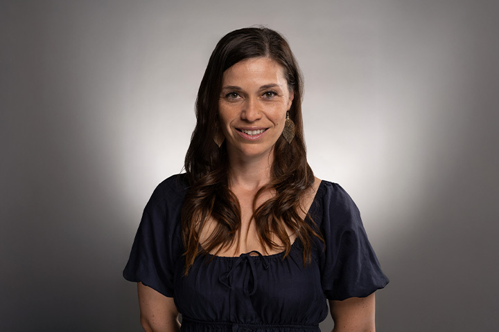 Sabine Duval with long, wavy brown hair wearing a dark blue blouse stands against a plain grey background.