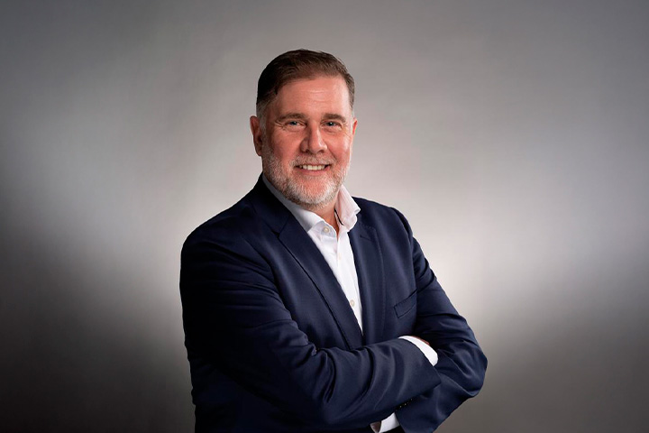 David Hickson, wearing a dark blue suit and white dress shirt standing with arms crossed against a gradient grey background. 