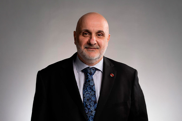 Joe Turnaturi, a bald man wearing a black suit, white shirt, and a blue patterned tie. The background is plain and grey.