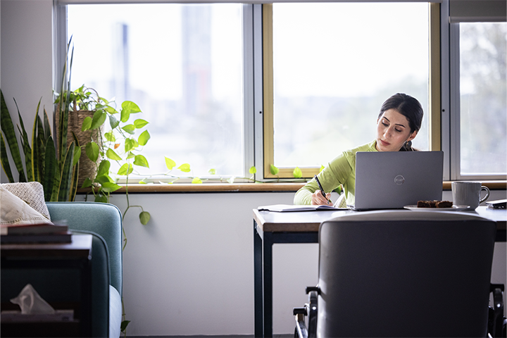 A woman sits at a desk in a bright, modern room, working on a laptop and taking notes by hand. Green plants and a coffee cup sit nearby, creating a calm and focused workspace.