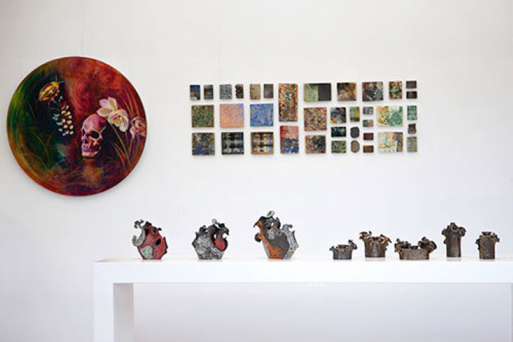 An assortment of small art works hang are exhibited on a white wall. In front there is a thin, long table on which ceramic work and plants are displayed.