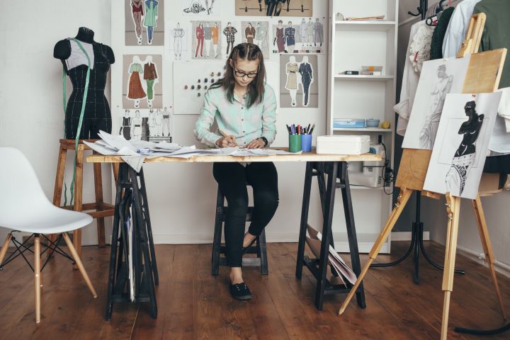 A woman dressed in a shirt and trousers sits at a desk working on some drawings. On the wall behind her are fashion sketches and designs. There is a mannequin with measuring tape draped over it to her right, and an easel with designs to her left.