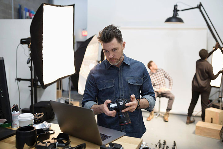 A man in a denim shirt and slicked back brown hair is holding a digital camera while looking at a laptop screen in a photography studio. There are people working behind him. One is seated in front of a white screen while the other adjusts overhead studio lighting.
