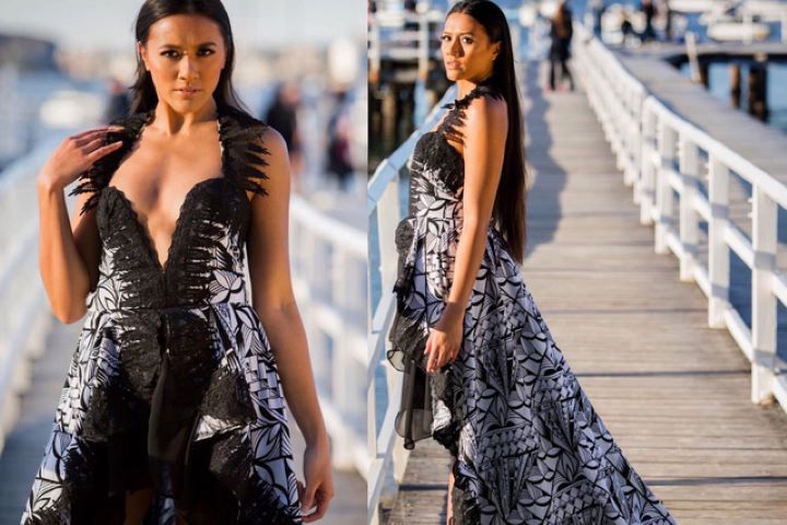 A model showcases a striking long, black-and-white dress with intricate patterns while posing on a pier. The dress features a plunging neckline and an asymmetrical hem, complemented by a flowing silhouette. There are blurred figure in the background further down the pier enjoying the clear sunny day.