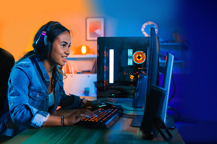 A young woman with dark hair is sitting in a dimly lit room at her desk. She is wearing headphones and engaged in a gaming activity on a computer screen with a smile on her face. 