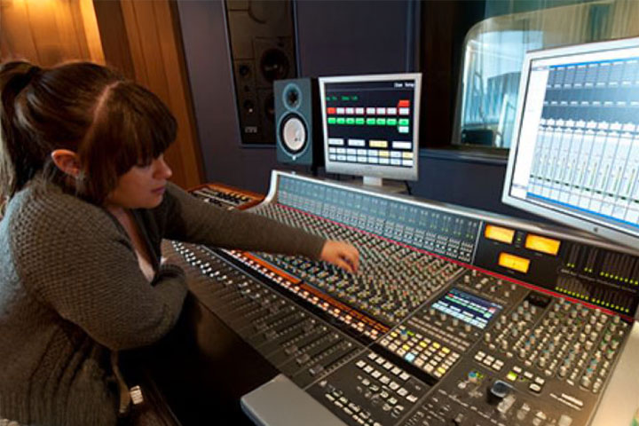A woman is working in a professional recording studio at an audio mixing console. She has multiple screens in front of her displaying audio waveforms as she adjusts sound levels on a large soundboard.