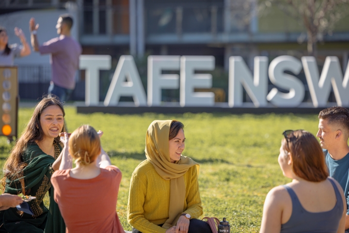 A group of students sit on the grass, talking and relaxing near a large "TAFE NSW" sign in the background.