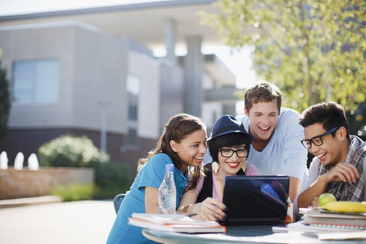 Four students sitting outside are gathered around a laptop. They’re smiling and looking at the screen.