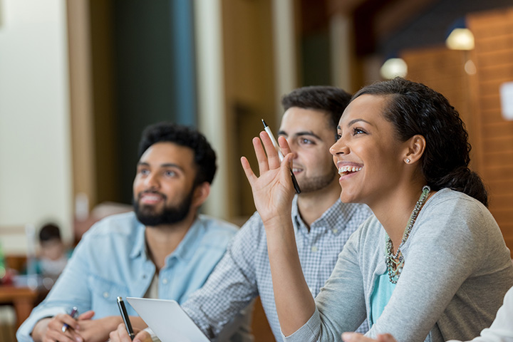 A group of students are seated at a table, with one person actively raising their hand to speak or ask a question.