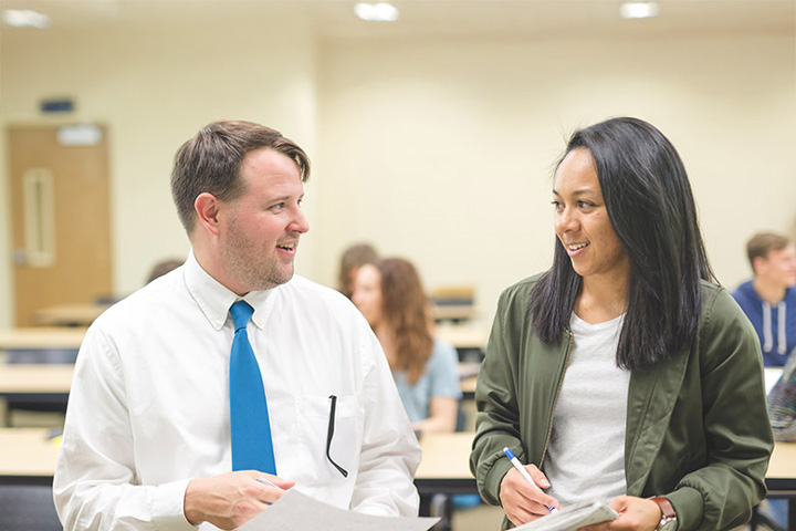 A man and woman engage in conversation in a classroom setting, surrounded by other students and desks.