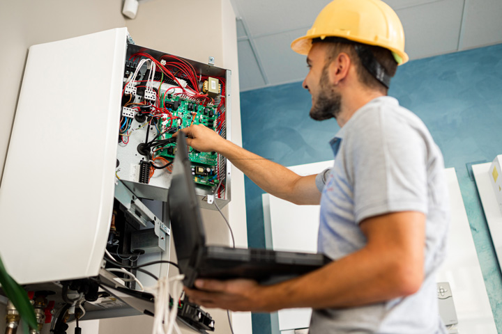 An electrician wearing a yellow hard hat works on an electrical panel, inspecting the wiring while holding a laptop for diagnostics.