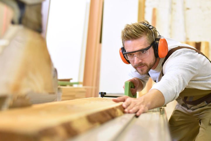 A man wearing safety glasses and ear muffs is bent over, looking along a plank of timber. He’s reaching along the wood with one hand, and in the other, he’s holding a hand tool. 