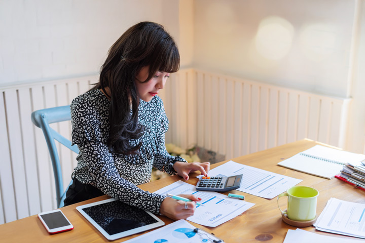 A young woman sits at a desk with papers and screens out. She is paying an invoice while using a calculator.