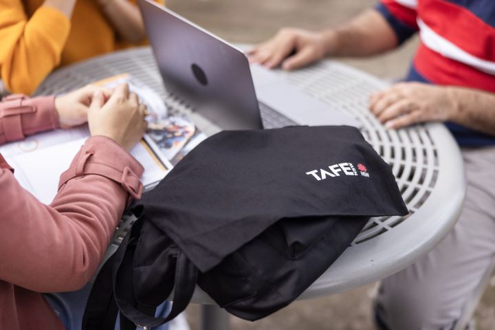 A TAFE NSW-branded bag on a table, with two people sitting nearby, a laptop, and documents in view.