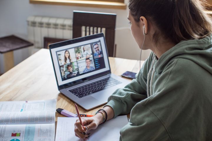 A young woman attends a meeting via Microsoft Teams. She has her headphones connected, a textbook open in front of her and is poised to take notes in her notepad. She wears sage green hoodie and has her hair in a high ponytail. 