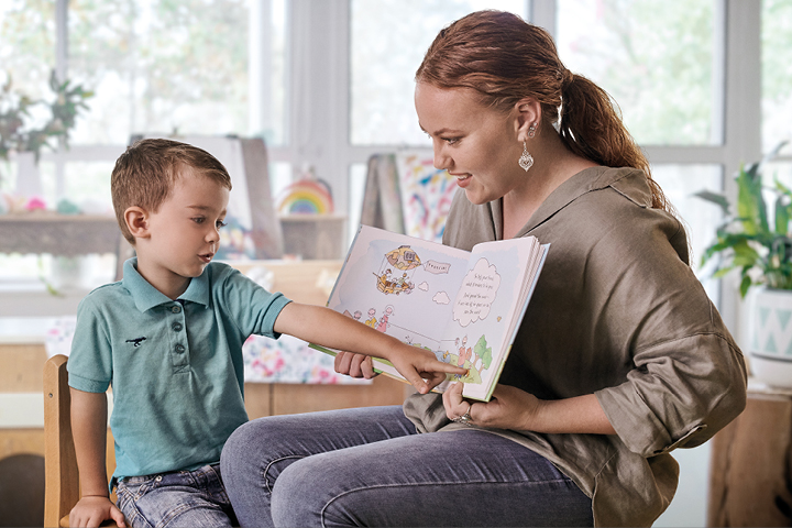 A young, smiling female early childhood educator with a preschool-aged child, holding a colourful picture book open in their laps. The child points at the page while the teacher looks on attentively, ready to help sound out words or answer questions.
