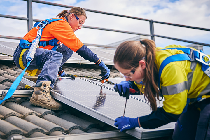 Two women in safety harnesses work together to install solar panels on a sunny rooftop.
